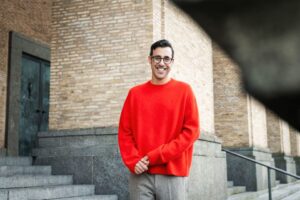 Image of a man standing in front of a brick building, he's wearing glasses and a bright red jumper. This is Ash Mann.